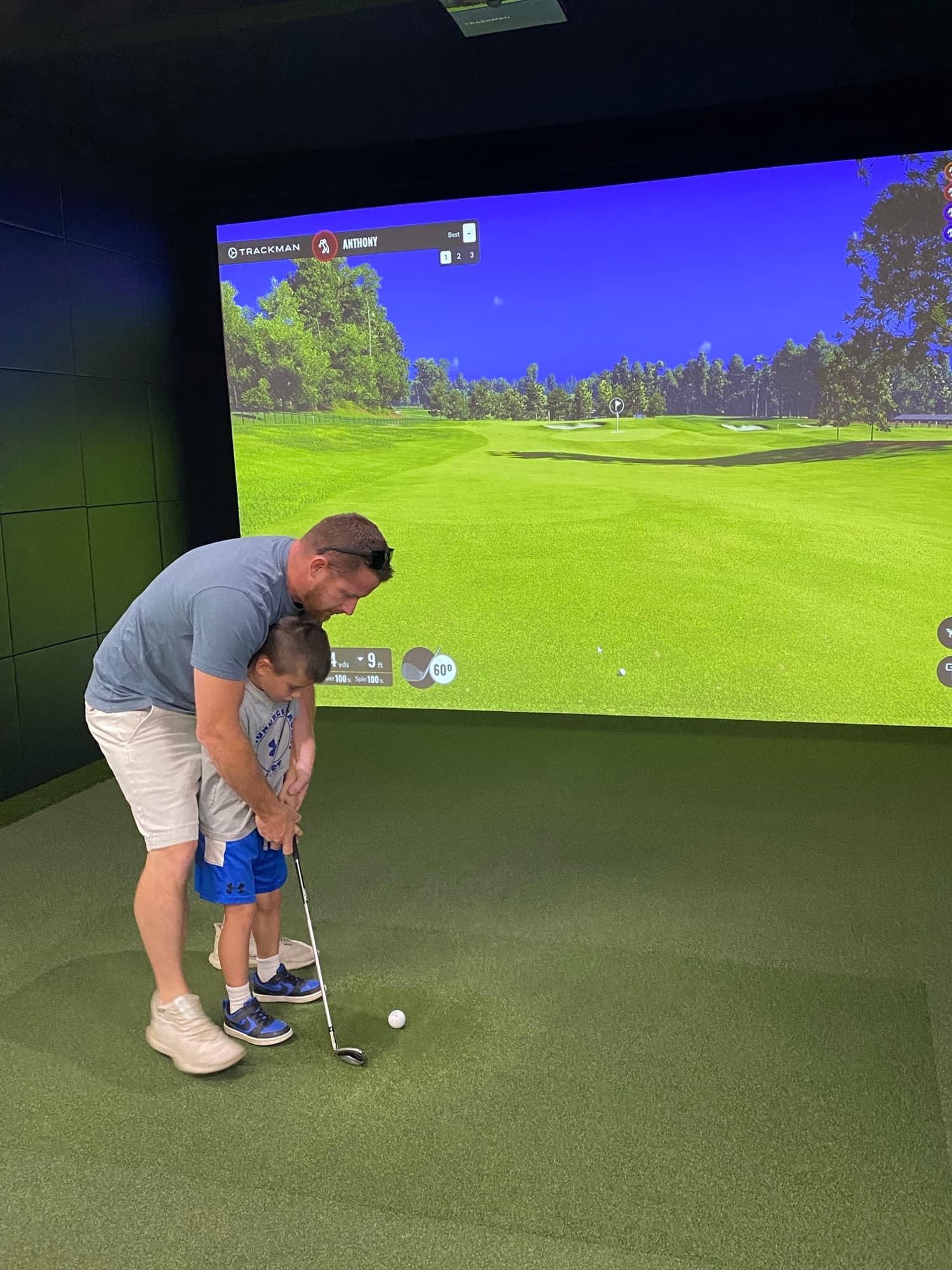 A parent helping a child with their swing in a simulator bay at Unbound Golf